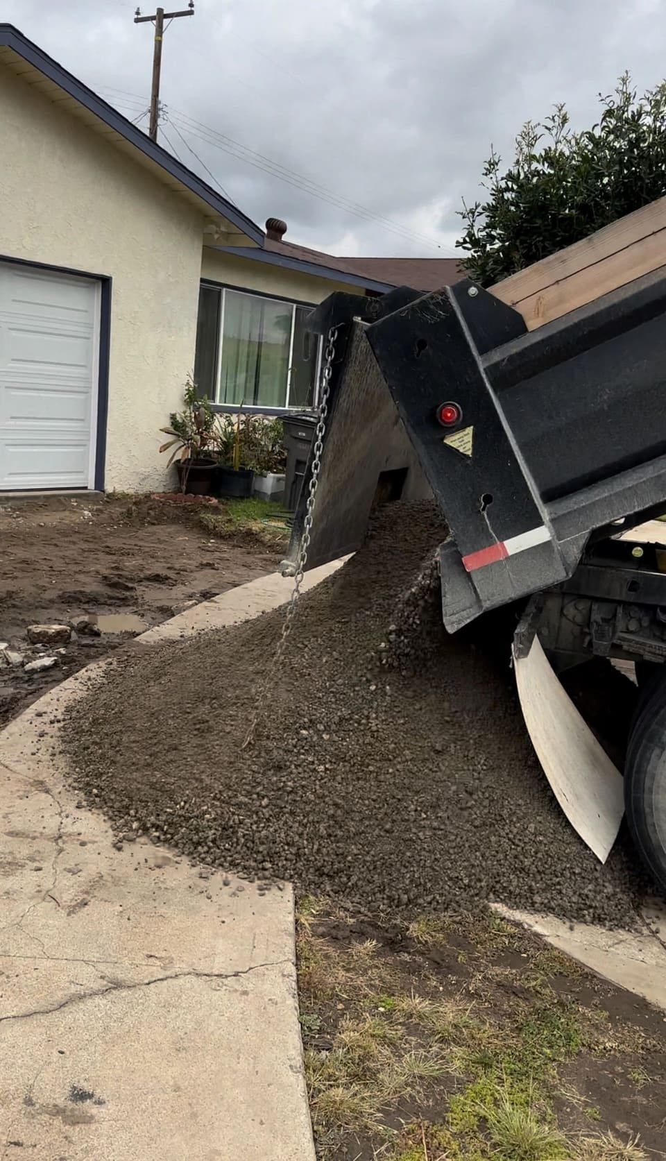 A dump truck with its bed raised pours dark gray crushed rock onto a concrete driveway in front of a house.