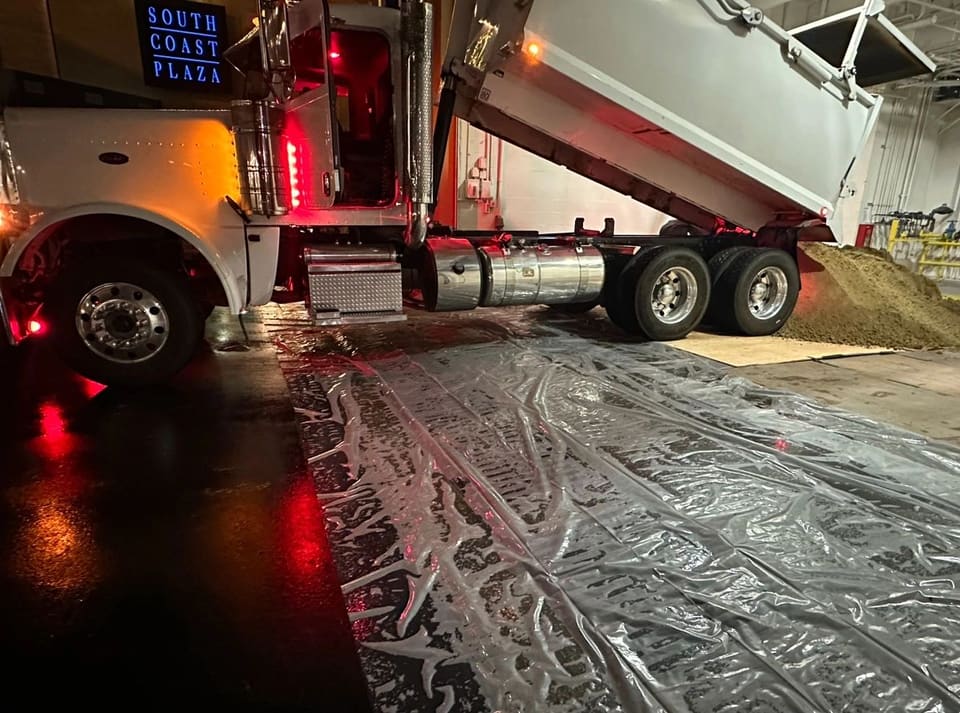 A white dump truck with its bed raised dumps tan material onto plastic sheeting in a covered lot at night.