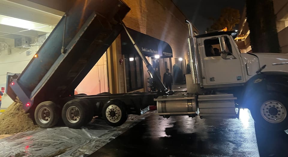 A dump truck with its bed raised tips tan material onto plastic sheeting on wet pavement at night.
