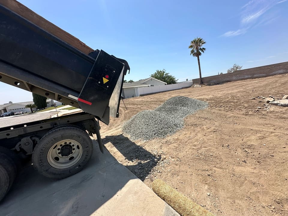 A dump truck with its bed raised has dropped a pile of gray crushed rock on bare dirt beside a concrete slab.