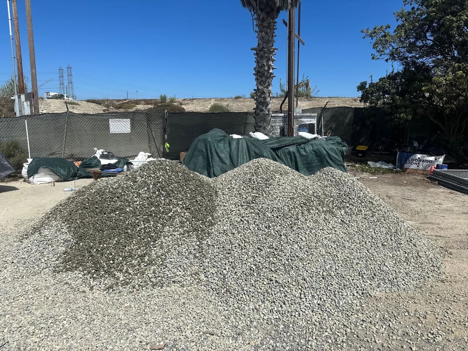 Two piles of gray crushed rock sit on bare dirt in a fenced yard with green tarps and a palm tree behind them.