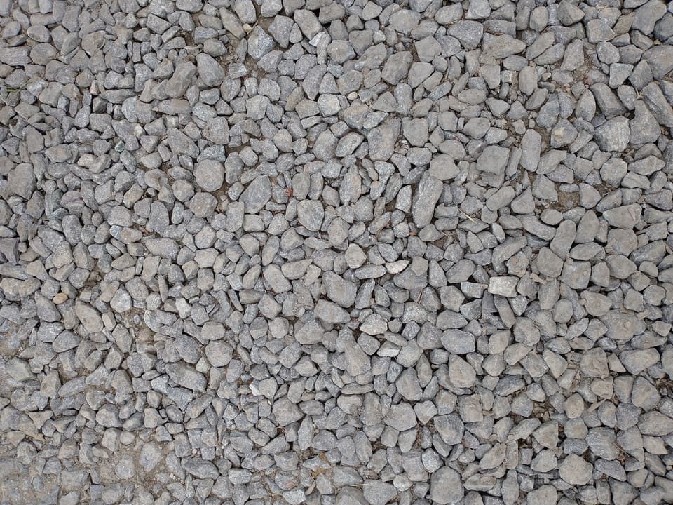 Top-down close-up of gray angular crushed rock in mixed sizes packed tightly together, with darker fines and dust filling the gaps between the pieces.