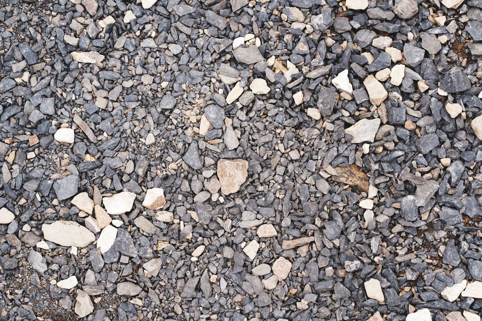Close-up of a gravel surface made of dark gray angular crushed stone in mixed sizes with scattered cream-colored pieces and fine grit between them.
