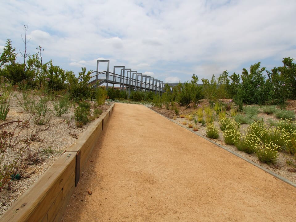 A straight, compacted tan sandy gravel path edged with timber beams runs between planted beds toward a raised metal walkway under a cloudy sky.