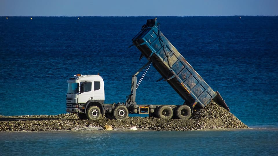 A white-cab dump truck with its bed raised tips gravel onto a narrow spit of stone jutting into deep blue water.