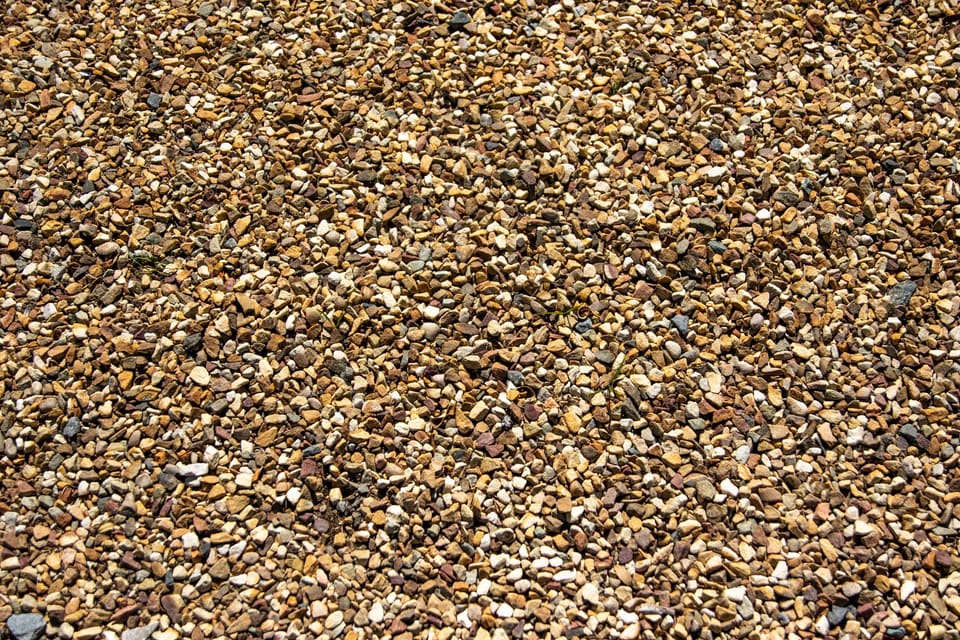 A sunlit close-up of small tan, brown and cream pea-sized gravel covering the ground with a few grass blades poking through.