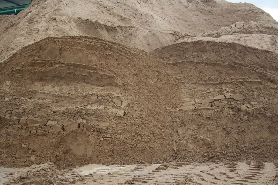A tall stockpile of damp tan washed sand with a loader-cut face showing fine grain and cracked lumps, with tire tracks in the foreground.