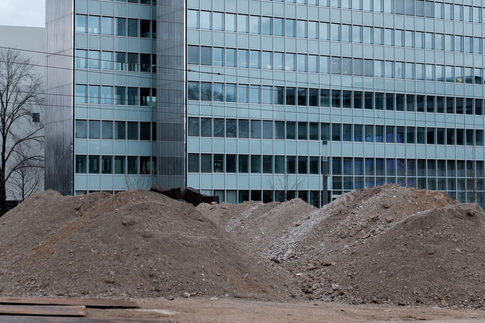 Heaps of grey-brown excavated earth mixed with rubble fragments on a bare lot in front of a multi-story glass office building, with bare trees and no people or machinery.