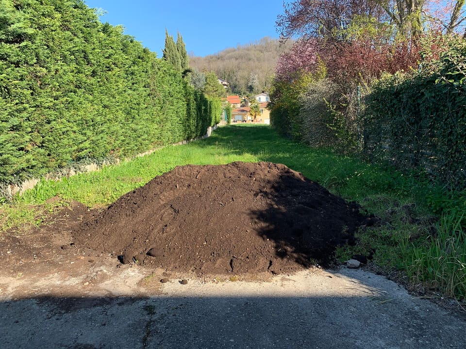 A rounded pile of dark brown, crumbly topsoil dumped on a concrete apron at the end of a grassy lane between tall hedges.