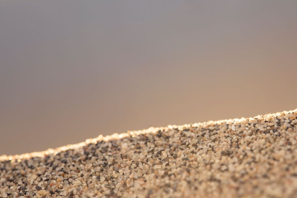 Coarse sand grains in mixed tan, gray, white and black tones piled on a slope in sharp focus across the lower half of the frame, with a plain blurred gray-tan background above.
